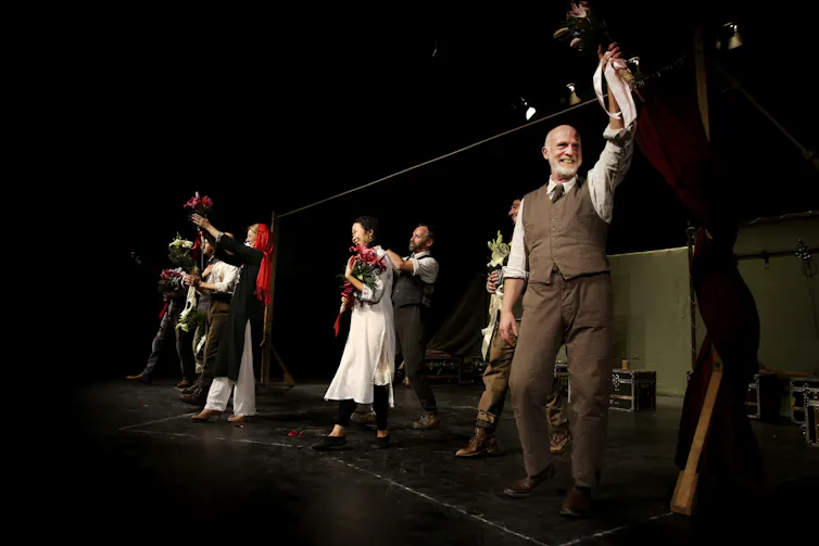 Actors on a stage, some holding flowers.