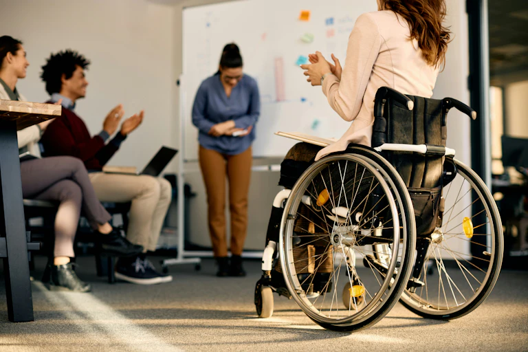 A person in front of a whiteboard bows while other people, including someone sitting in a wheelchair, clap for them