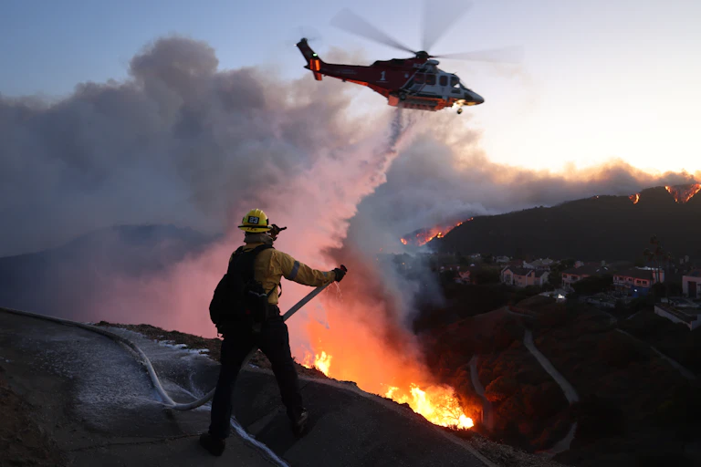 A firefighter trains a hose on a fire in a canyon with house in the background, while a helicopter drops water on the fire.