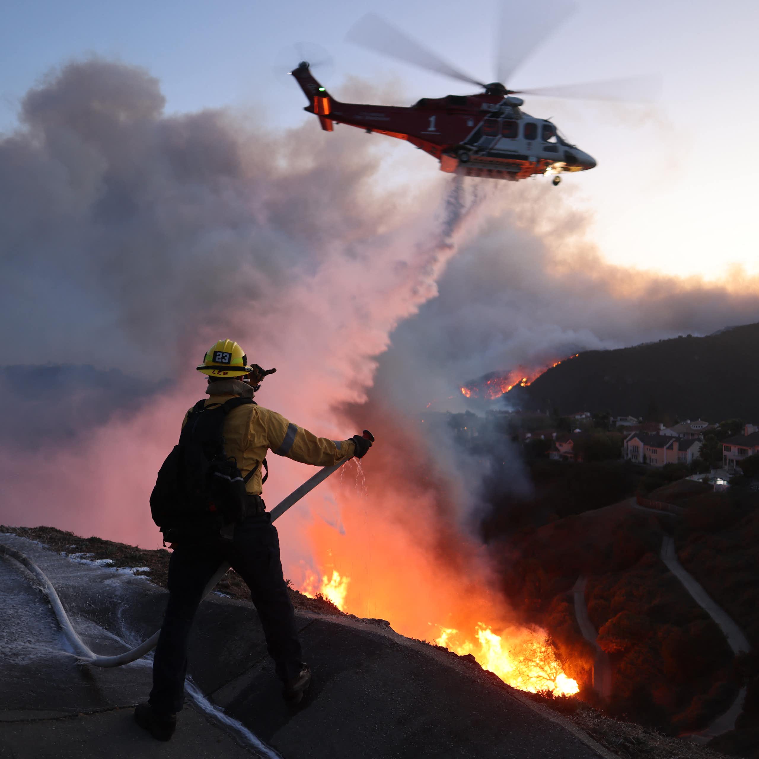 A firefighter trains a hose on a fire in a canyon with house in the background, while a helicopter drops water on the fire.