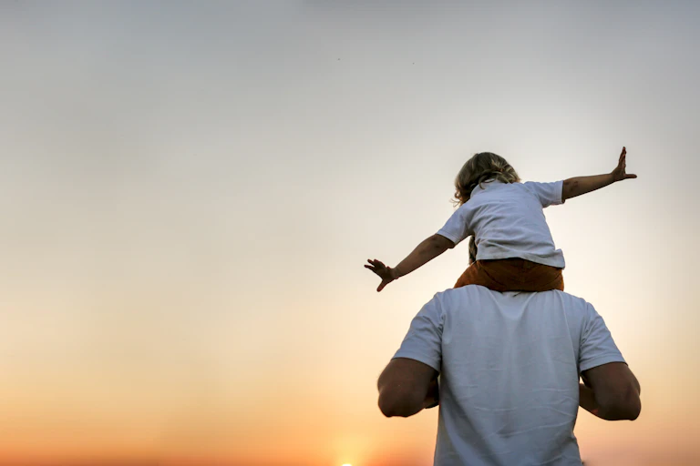A man holds a young child on his shoulders. The child has their arms outstretched