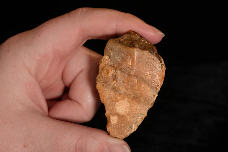 A hand in close-up holding an amber coloured chipped stone against a black background.
