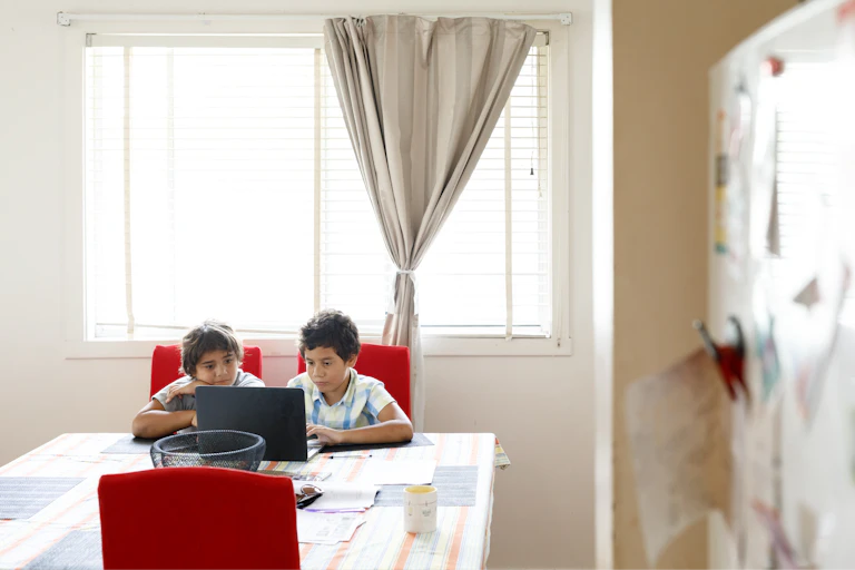 Two young Indigenous boys sit at a kitchen table and look at a laptop