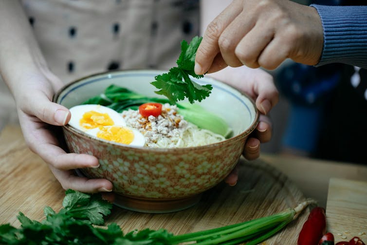 People add eggs and garnishes to a bowl of noodles