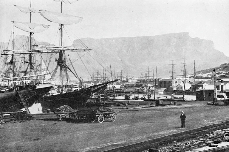 Old photo of sailing ships at harbour, with a mountain in the background and a cart in the foreground