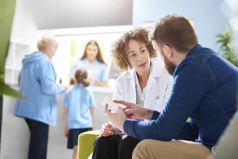 Pharmacist wearing white coat consulting male patient in GP clinic waiting room
