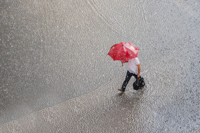 un homme marche avec un parapluie rouge dans une pluie torrentielle. L'image est vue d'en haut.