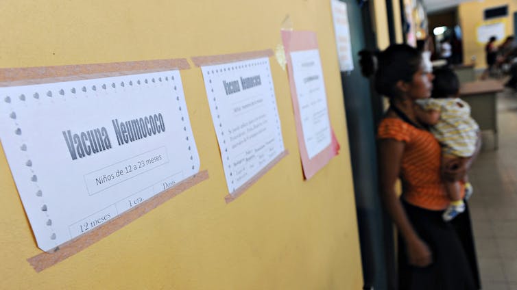 A woman holding an infant stands in a hallway with papers affixed to the walls.