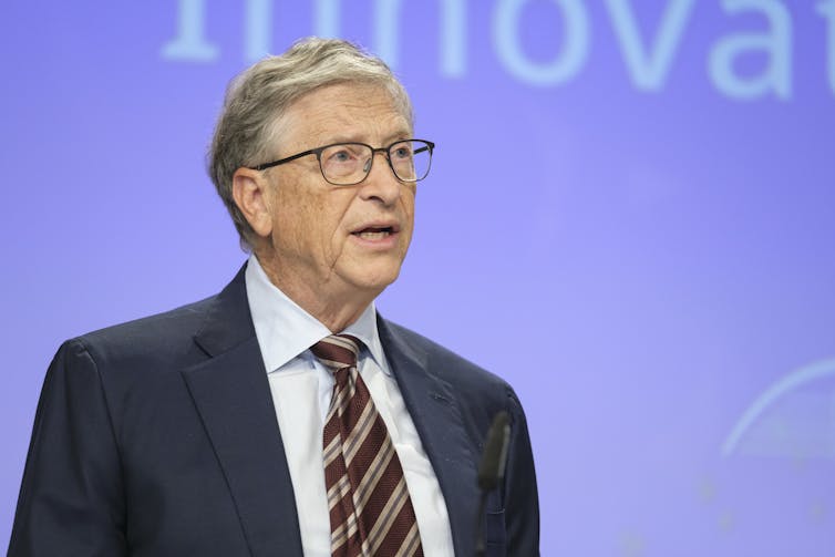Bill Gates in a suit and tie stands against a purple background.