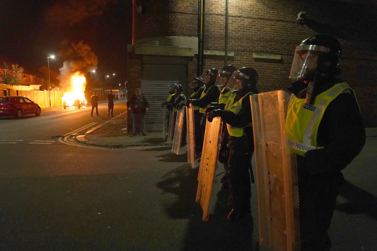 A row of police with riot shields stand in the darkness, in the background a car burns