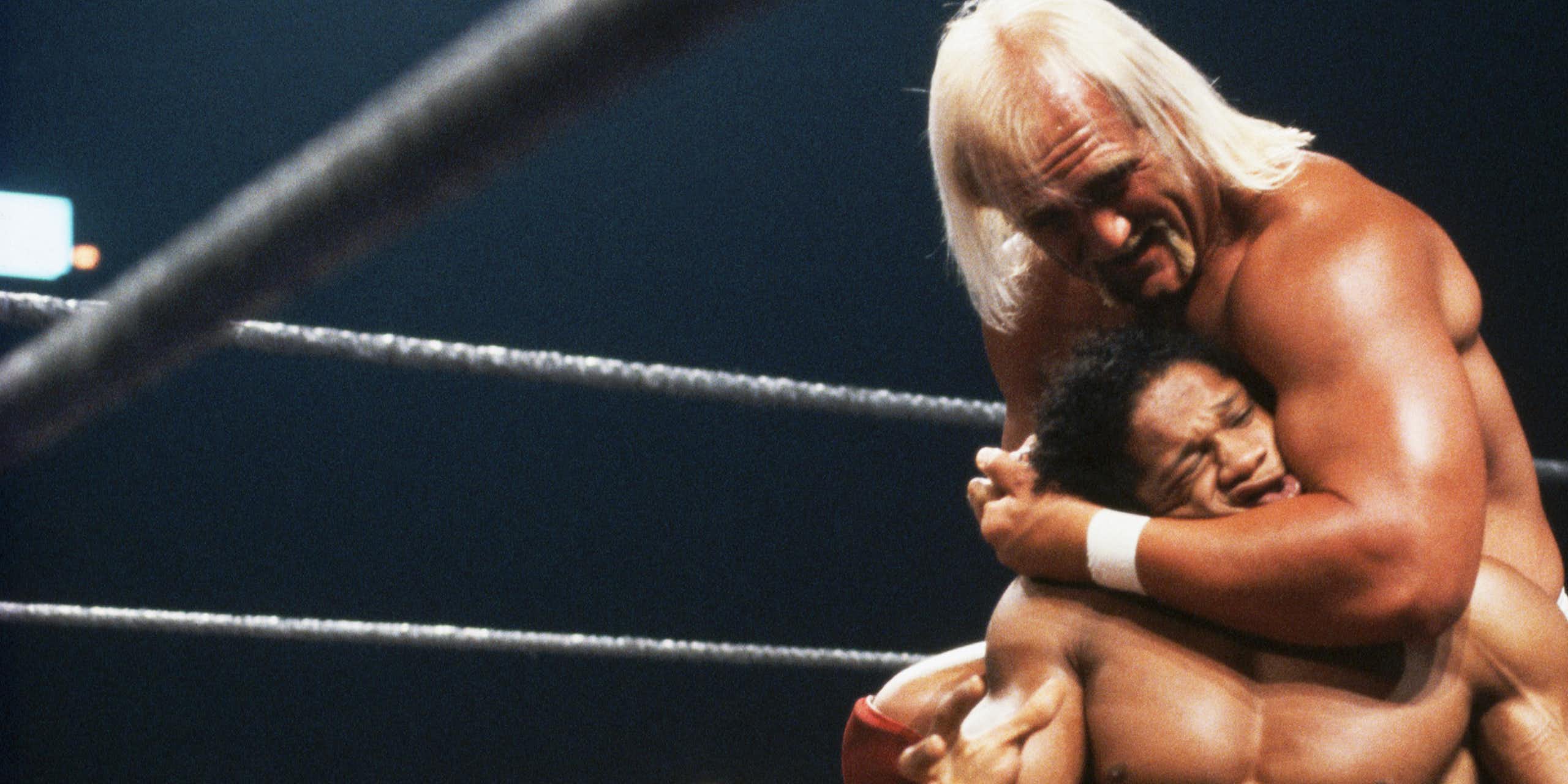 Muscular man with long, blond hair puts another man in a headlock during a wrestling match.