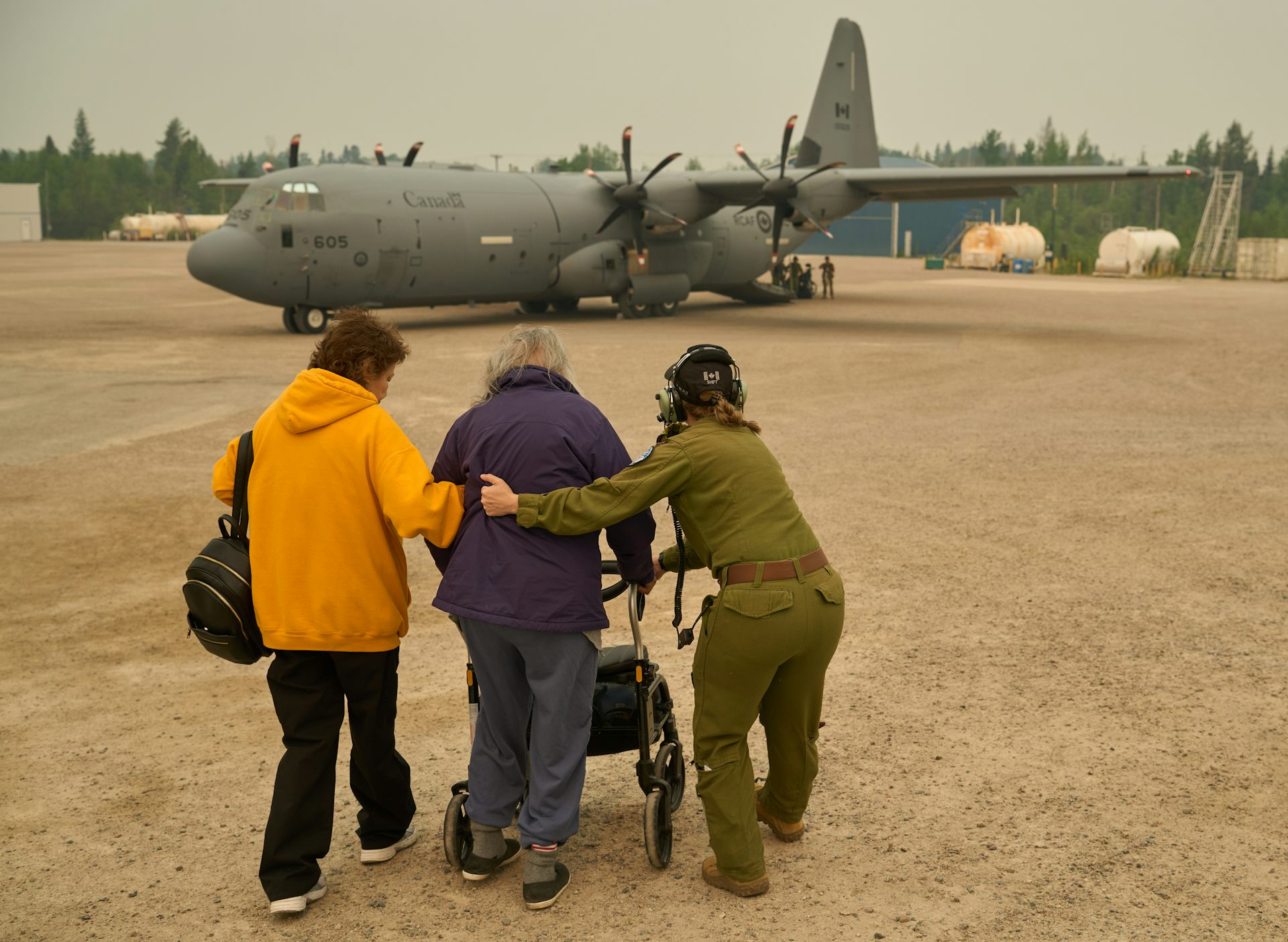 A woman in green military fatigues guides two older women, one using a walker, toward a grey military plane on the tarmac 