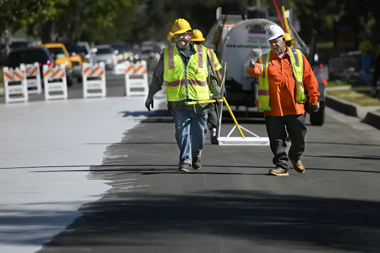Two men in reflective construction vests paint a stretch of road a light color. The difference between the dark and light is evident.