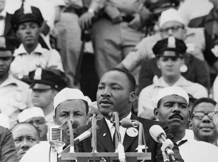 Martin Luther King Jr speaks into microphones with people in the background.