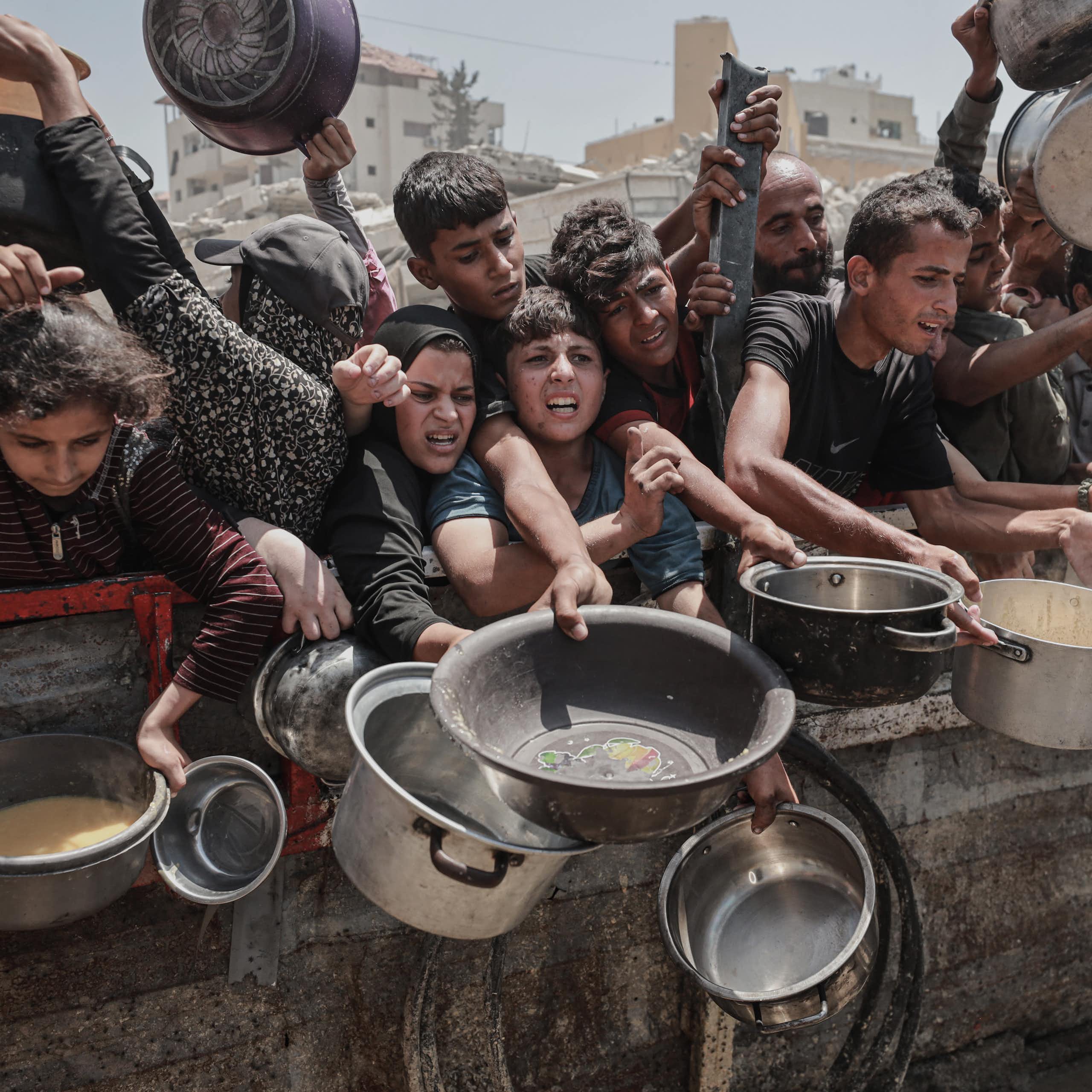 Children and adults crowded against a wall, holding out containers to get food.