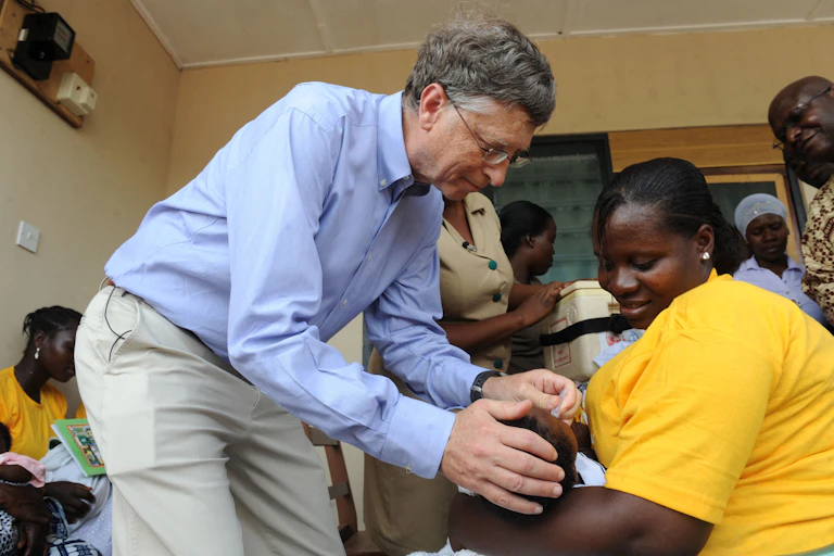 Bill Gates leans over a baby held by a woman in a bright yellow shirt.