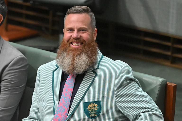 A bearded man in an Olympic green striped jacker grinning up from a seat in Parliament House, Canberra.