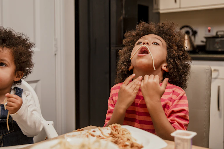 A young boy puts spaghetti over his mouth like a moustache at the dinner table.