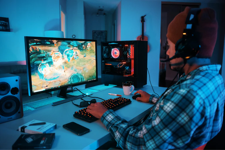 A young man sits at a desk playing a video game on a desktop computer. He is wearing headphones
