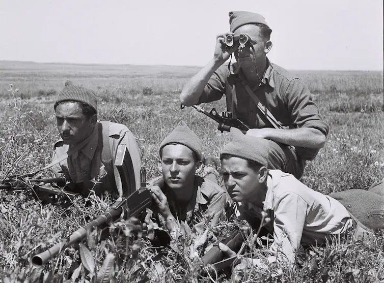 Four men in military uniform, one is spotting with binoculars, another has a rifle.