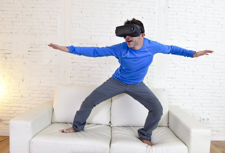 Man standing on sofa in surfing pose wearing VR headset.