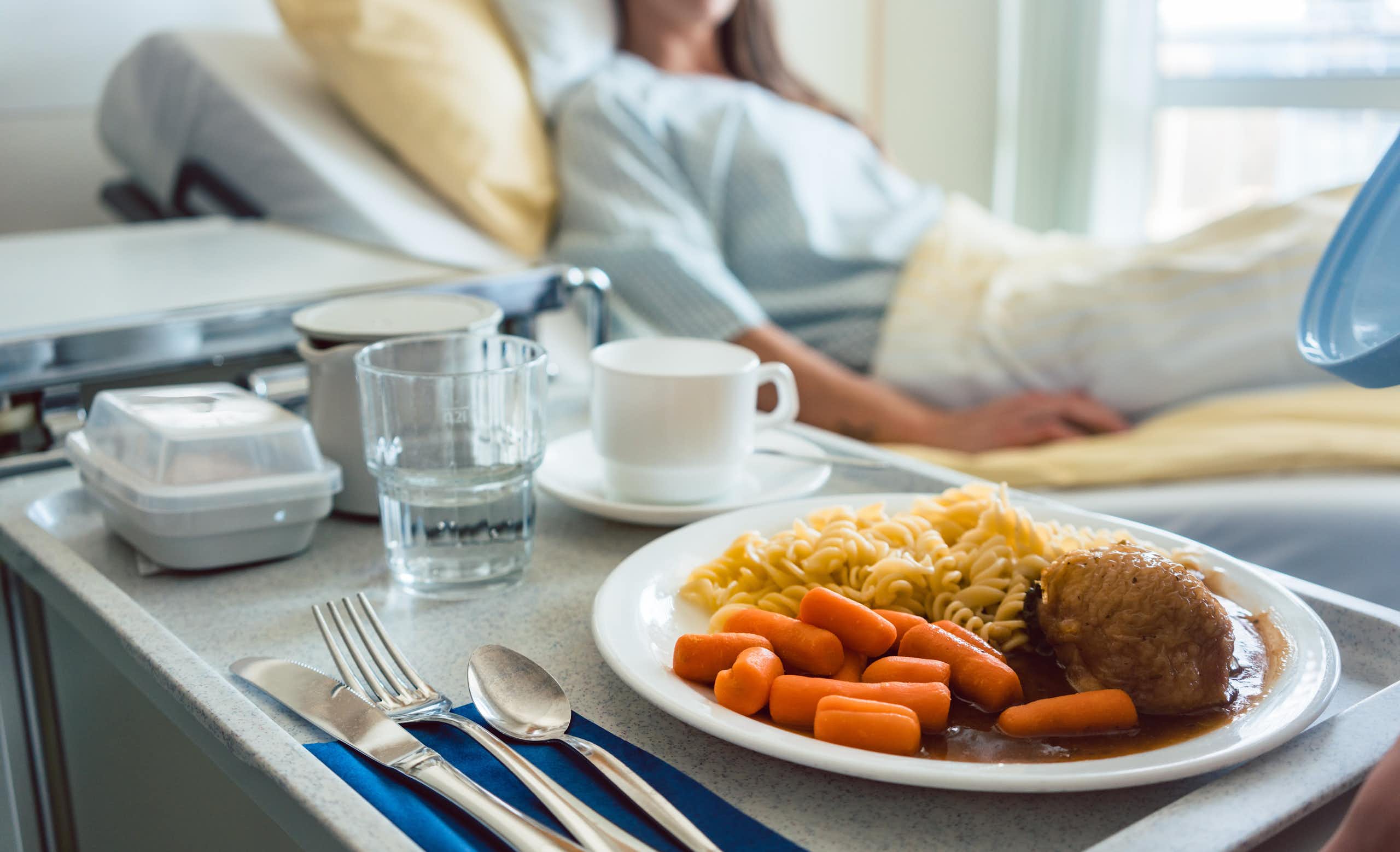 Comida sobre una bandeja sobre un carrito con una mujer acostada en una cama de hospital de fondo