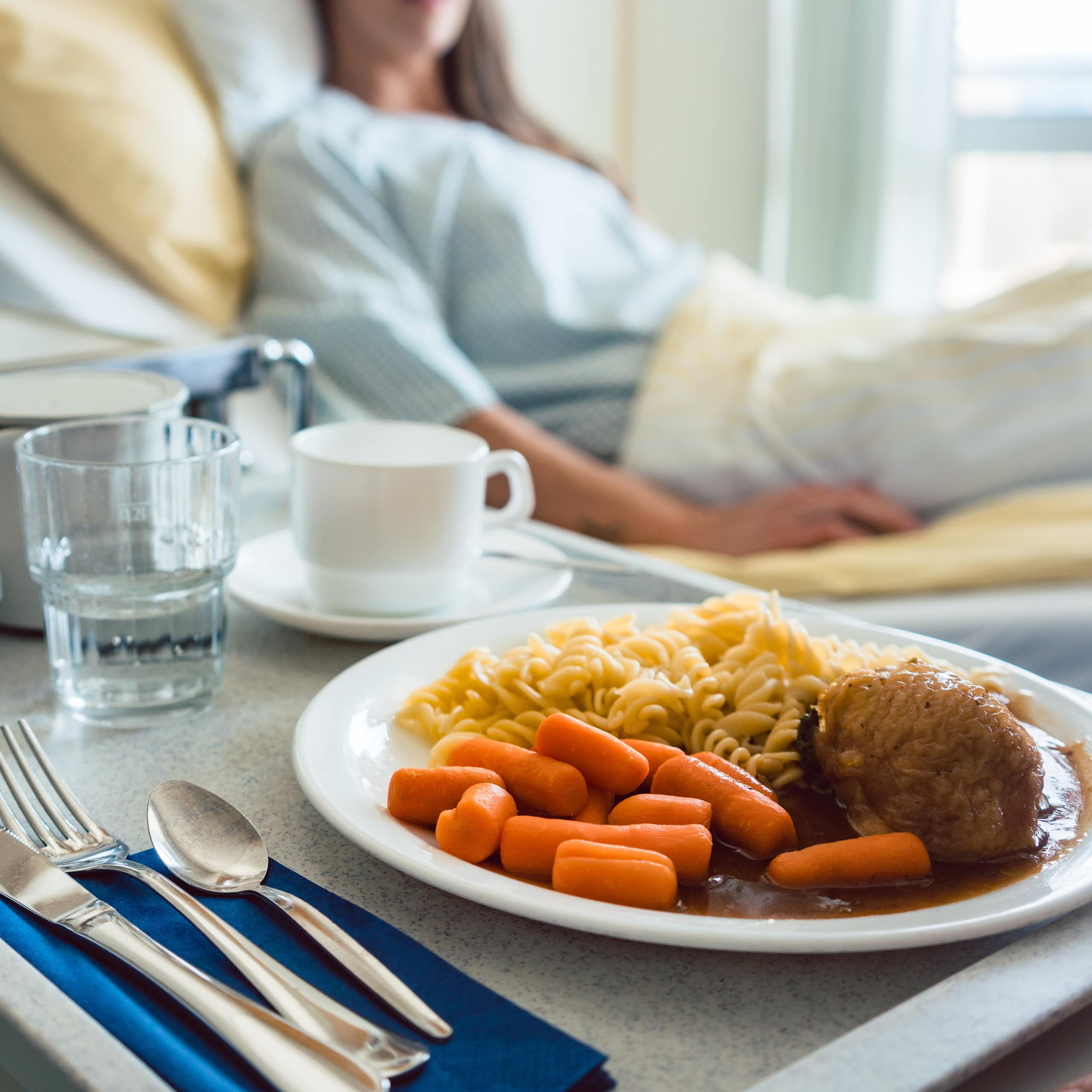 Comida sobre una bandeja sobre un carrito con una mujer acostada en una cama de hospital de fondo
