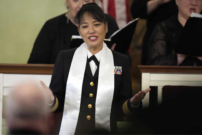 A woman, dressed in a black jacket with gold buttons and wearing a white sash around her neck, extends her arms as she sings.