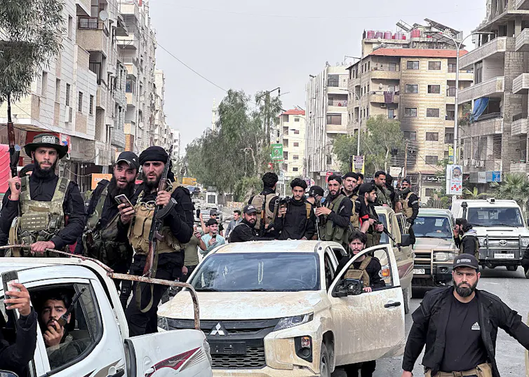 Two small white trucks with many soldiers standing on top of them drive through a wide city street.
