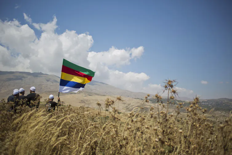 Four men in black clothing and white caps, seen from the back, hold a rainbow-striped flag as they stand in a vast field.