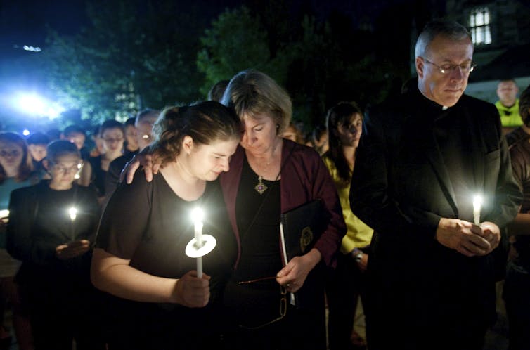 Several people stand mournfully while holding candles and one woman puts her hand around the shoulder of a younger woman in a consoling gesture.