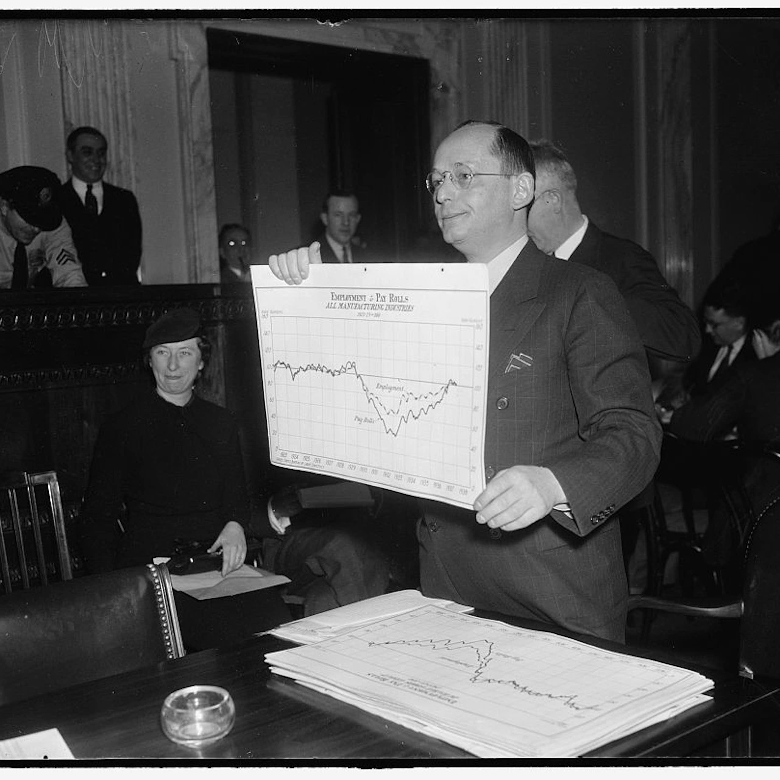 A man holds up a huge chart during a hearing in a black and white photo taken in the early or mid-20th century.