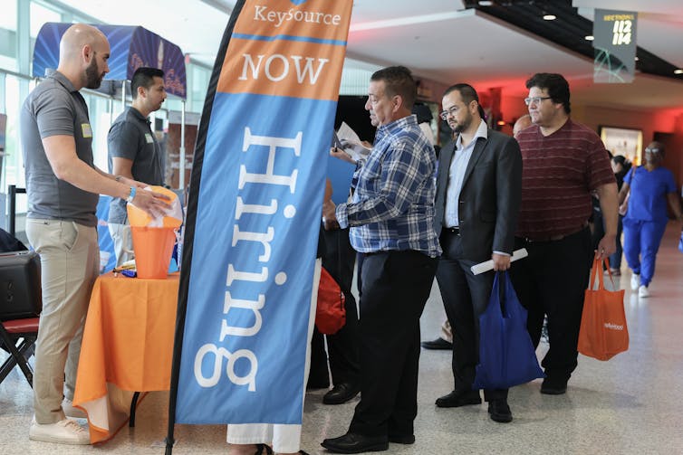 A group of men line up at a booth beside a banner that says 'now hiring.'