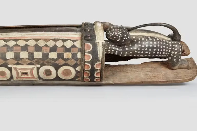 A carved drum with square and circular patterns and a lizard-humanlike creature pushing it.