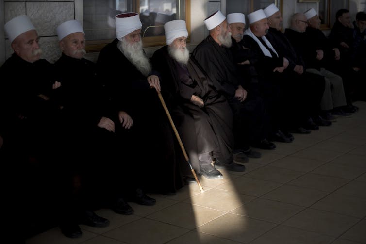 Men with flowing white beards, dressed in black robes and wearing white headdresses, are seated in a row.