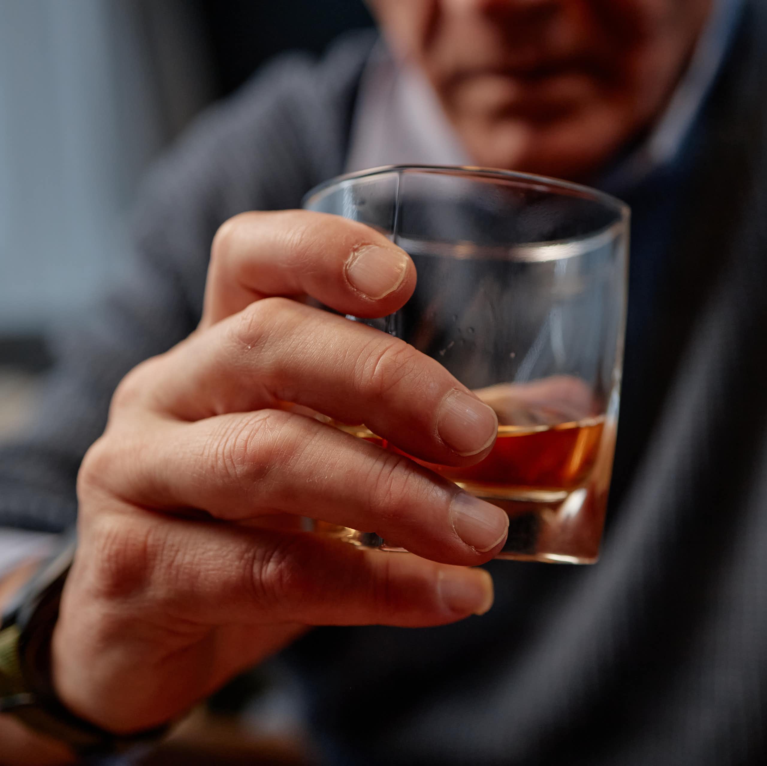 Gros plan sur la main d’un homme d’un certain âge tenant un verre qui semble contenir de l’alcool.