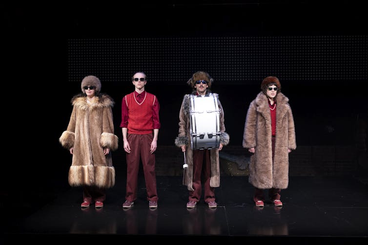Actors don Russian fur coats and sunglasses while standing on a stage.