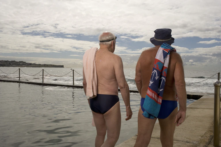 Two older men in speedoes walk with towels and goggles next to an sea pool.