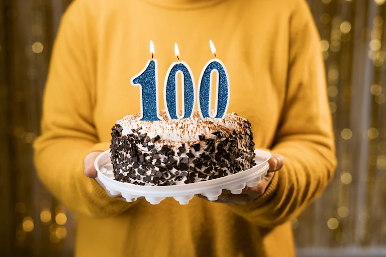 A person holds a cake that has lit candles in the shape of the number "100".