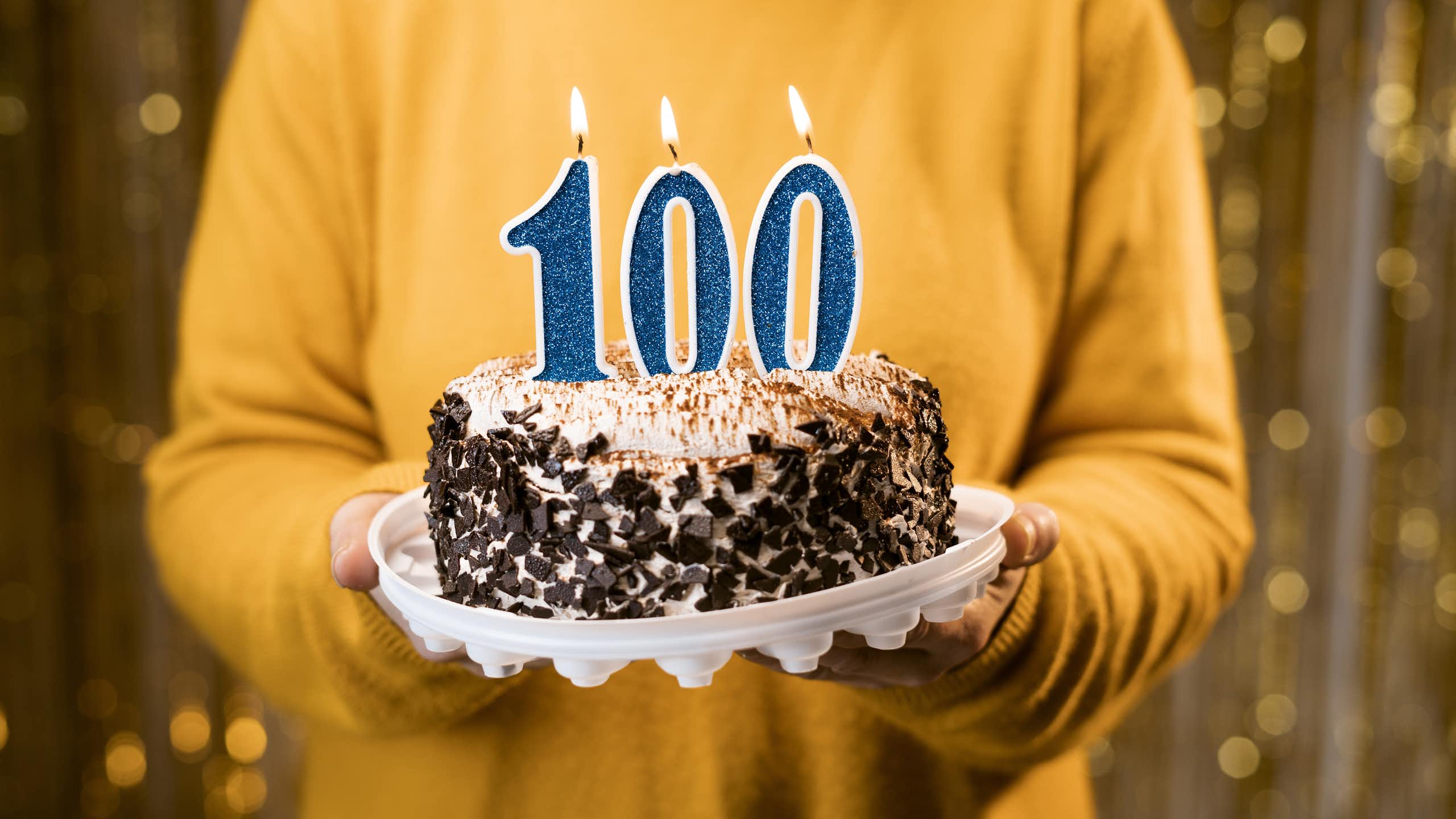 A person holds a cake that has lit candles in the shape of the number "100".