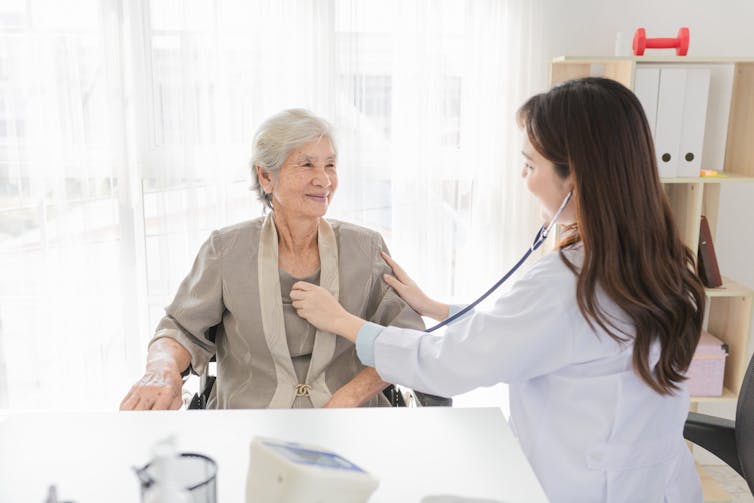 An elderly woman has her heart checked by a female doctor who is using a stethoscope.