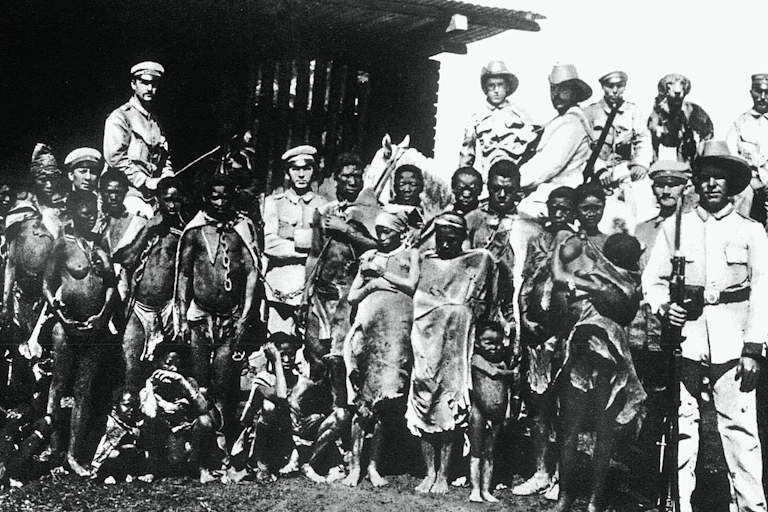 Una foto en blanco y negro muy antigua muestra a africanos casi desnudos encadenados entre sí, con soldados uniformados posando entre ellos.