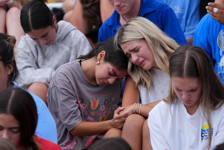 Adolescents sit in mourning, with two leaning on each other crying, at a memorial service.