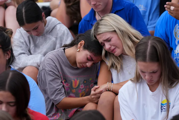 Adolescents sit in mourning, with two leaning on each other crying, at a memorial service.