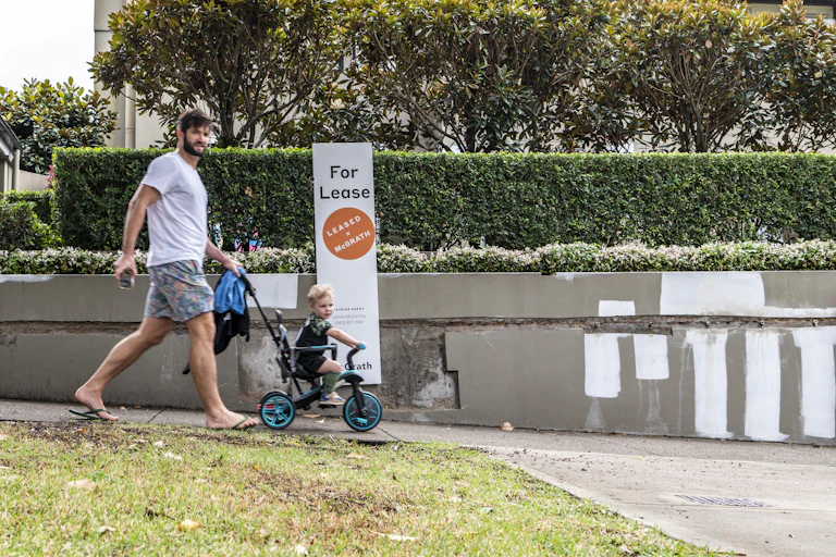 A man pushes a toddler on a bike outside a property with a 'for lease' sign