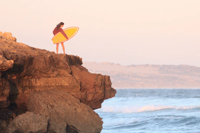 Girl clutching yellow surfboard stands on small cliff looking at ocean.