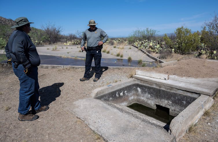 Two men stand beside a cement box. The landscape is dry around them.