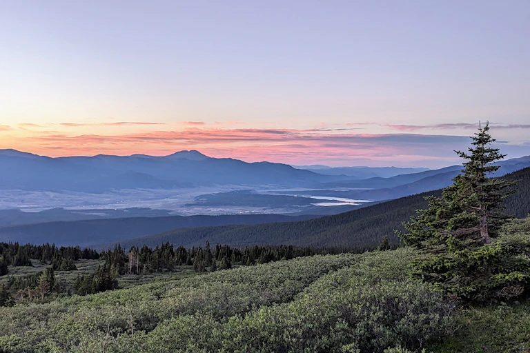 A magestic sunrise paints the mountainous sky with orange and purple clouds. A tree is in the foreground and bodies of water are visible in the distance.