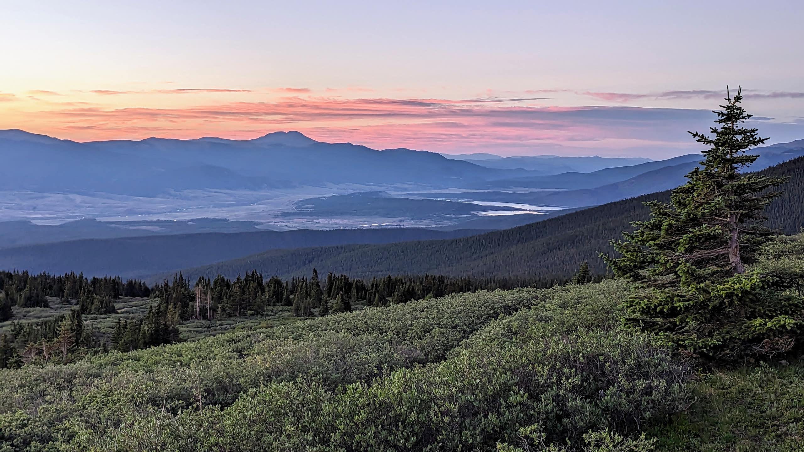 A magestic sunrise paints the mountainous sky with orange and purple clouds. A tree is in the foreground and bodies of water are visible in the distance.