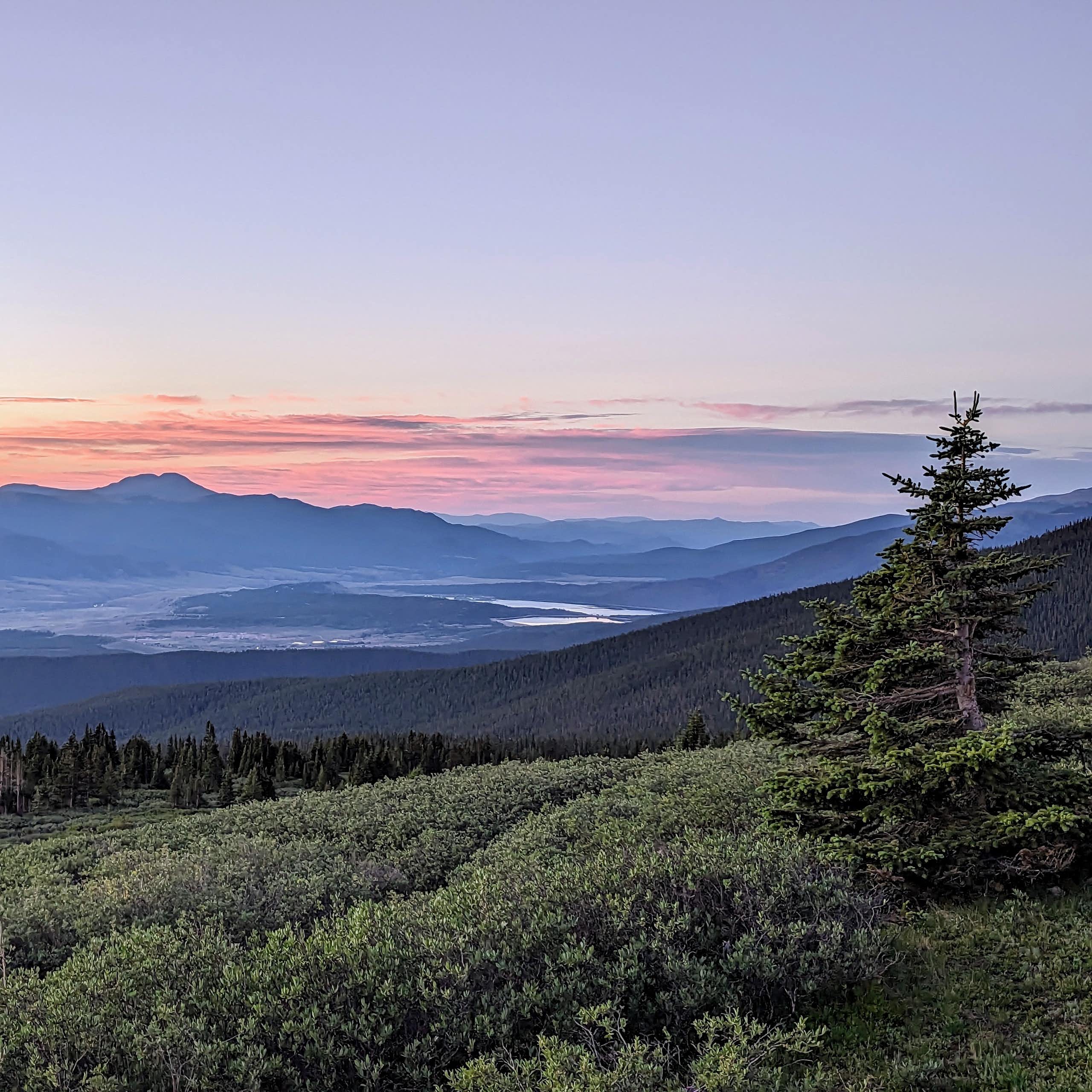 A magestic sunrise paints the mountainous sky with orange and purple clouds. A tree is in the foreground and bodies of water are visible in the distance.